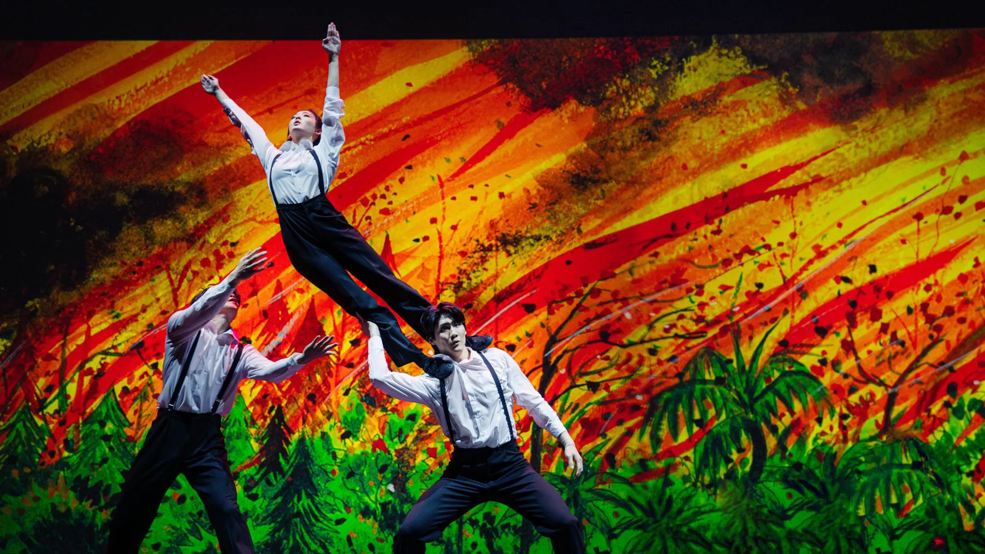 Three FOCASA Circus performers on stage in white shirts and black trousers, with one acrobat lifted mid-air against a vivid orange and green painted backdrop.
